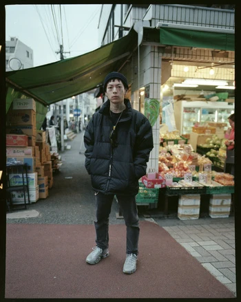 Portrait of a person with tattoos and a beanie in a black puffer jacket standing outside of a market stall on a city street.