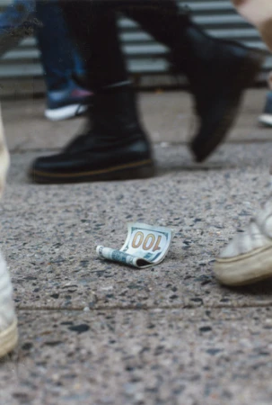 A one hundred dollar bill lies on a paved urban ground, with the blurred legs and feet of several people in the background.