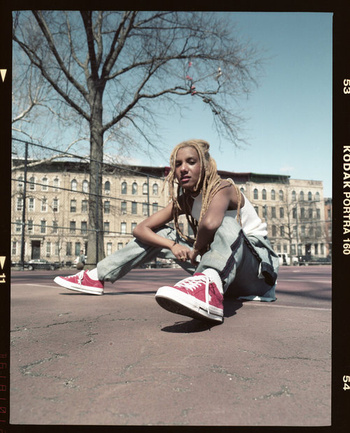Person with dreadlocks wearing a white tank top and red sneakers seated outdoors on asphalt with a building and tree behind.