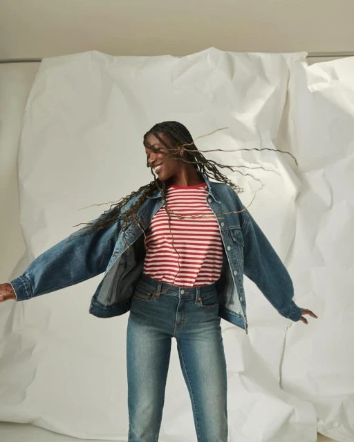 A joyful Black woman in jeans and a striped shirt smiles against a white backdrop, exuding casual style and denim fashion.