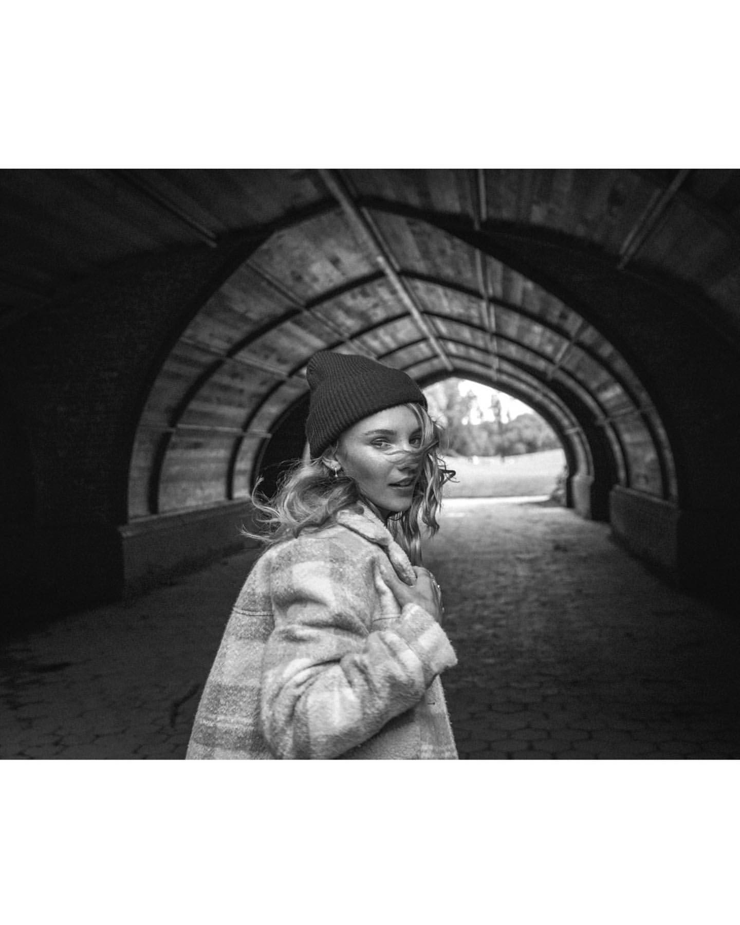 Black and white portrait of a woman in a tunnel wearing a beanie and plaid coat looking back over her shoulder at the camera.