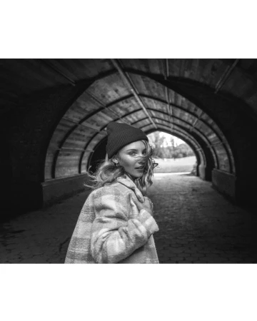 Black and white portrait of a woman in a tunnel wearing a beanie and plaid coat looking back over her shoulder at the camera.