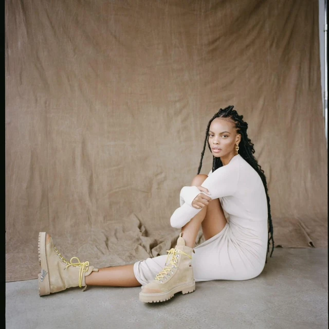An adult woman poses indoors, seated on the floor wearing a long-sleeve dress and tan-colored boots with yellow laces against a draped backdrop.