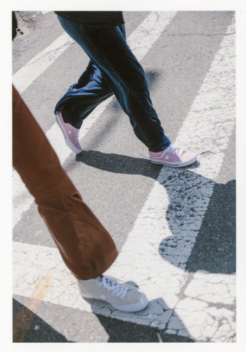 Two figures walk on a pedestrian crosswalk with light colored sneakers. The light is bright and there are stark shadows visible.