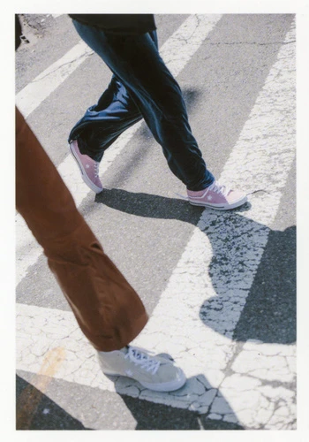 Two figures walk on a pedestrian crosswalk with light colored sneakers. The light is bright and there are stark shadows visible.
