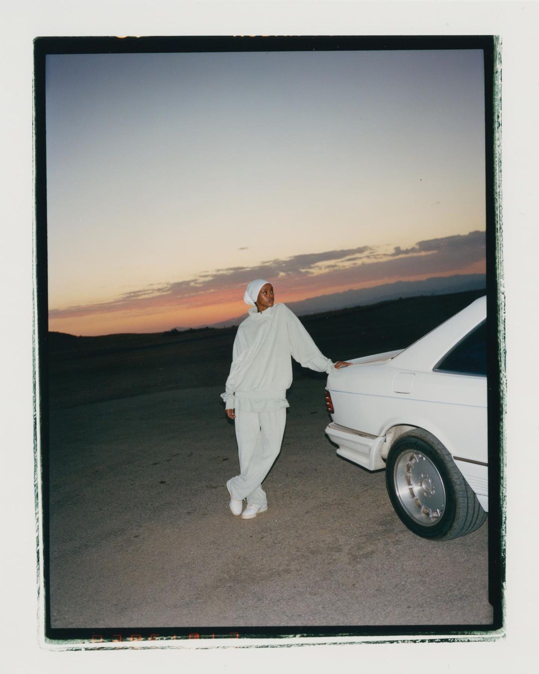 Adult poses in a light-coloured tracksuit beside a white classic car under a dramatic dusk sky in a minimal outdoor environment.