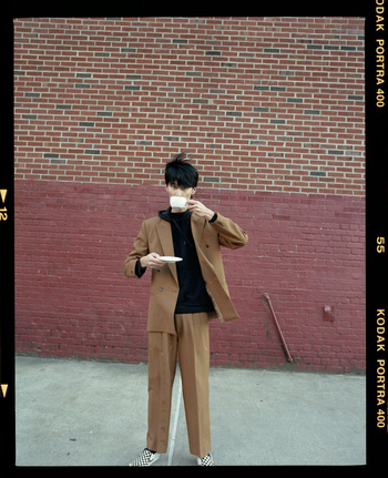 Young man in a brown suit and black hoodie stands against a brick wall and drinks from a teacup, posing in a film-framed photograph.