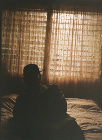Silhouetted adult sits on a bed with a view of a window with sheer curtains and a building in the background during the day.