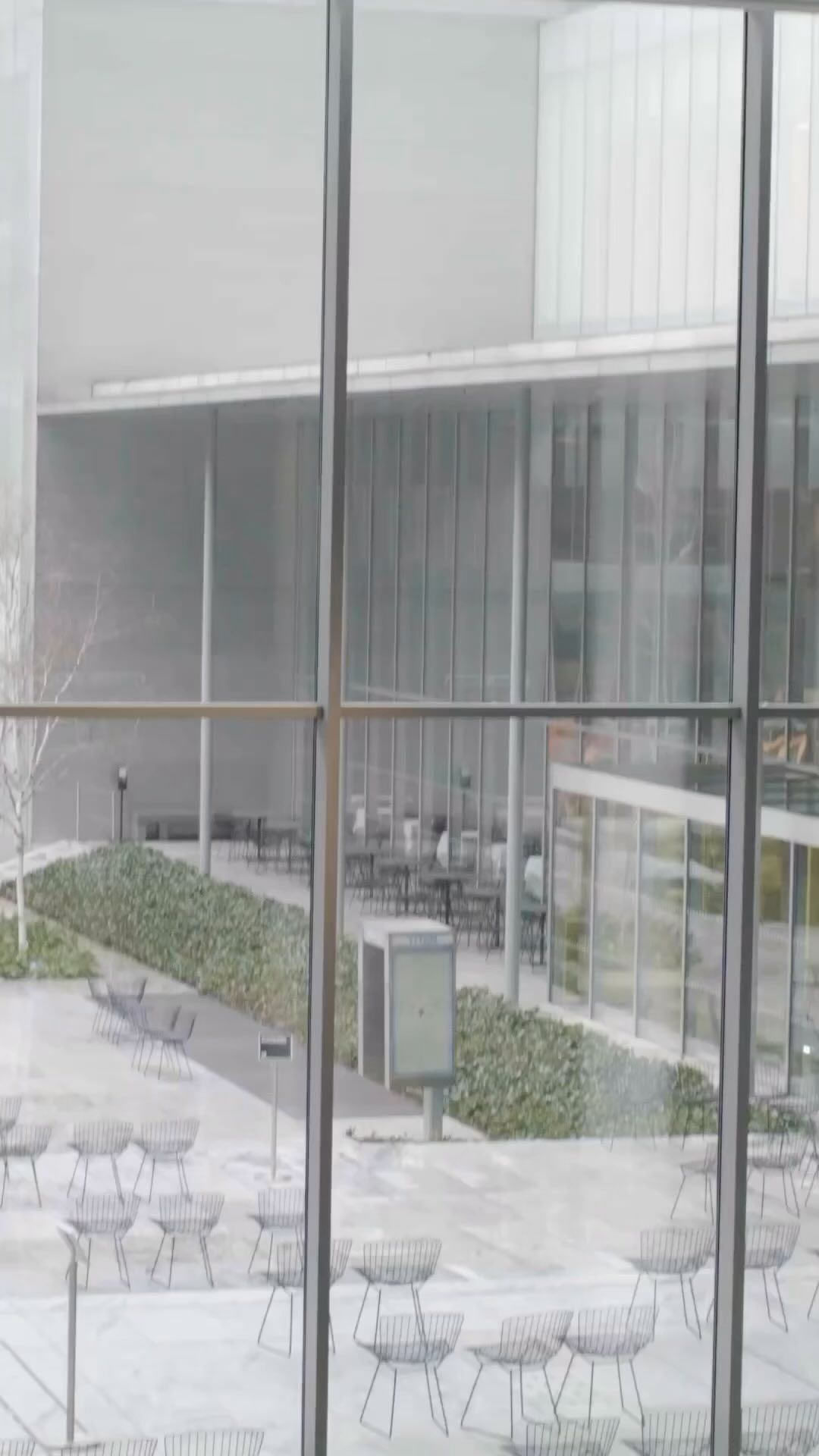 Outdoor courtyard view through a large window, featuring chairs and tables in a neutral-toned architectural setting with sparse vegetation.