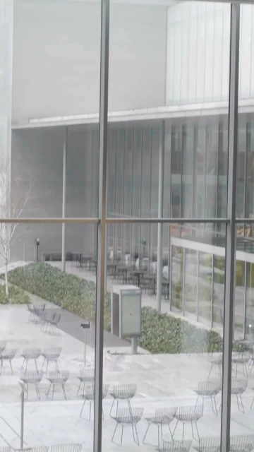 Outdoor courtyard view through a large window, featuring chairs and tables in a neutral-toned architectural setting with sparse vegetation.
