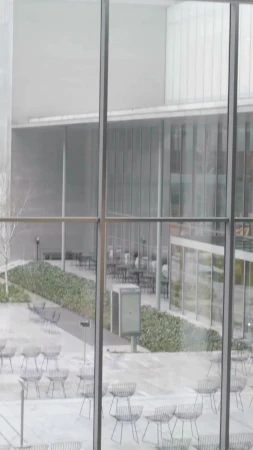 Outdoor courtyard view through a large window, featuring chairs and tables in a neutral-toned architectural setting with sparse vegetation.