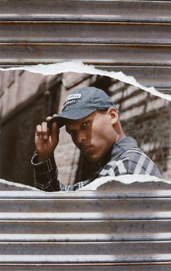 Portrait of an African-American man wearing a cap and plaid shirt, peeking through a torn metal barrier with a brick building background.