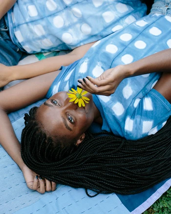 Eye-level shot of two women lying on a patterned blue quilt on grass, wearing matching tie-dye shirts, one holding a yellow flower.