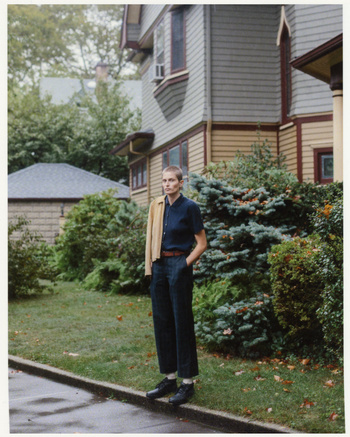 A woman with short hair poses in front of a two-story house on a cloudy day. She wears a collared shirt and jacket.