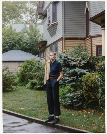 A woman with short hair poses in front of a two-story house on a cloudy day. She wears a collared shirt and jacket.