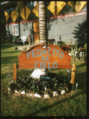 A wooden sign with 'Pedacito De Cielo' in blue lettering, surrounded by plants and set against a building and a palm tree.