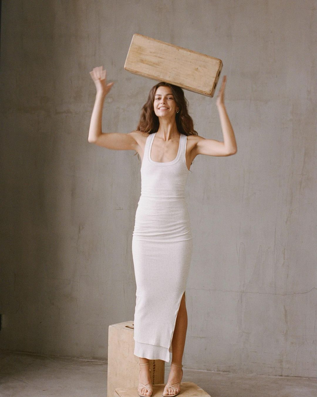 A woman in a white dress balances a rectangular wooden object on her head in a neutral indoor setting, smiling with arms raised.
