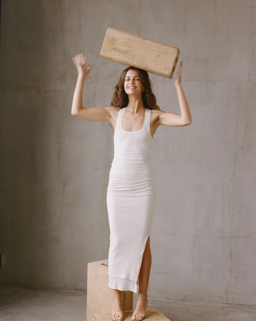 A woman in a white dress balances a rectangular wooden object on her head in a neutral indoor setting, smiling with arms raised.