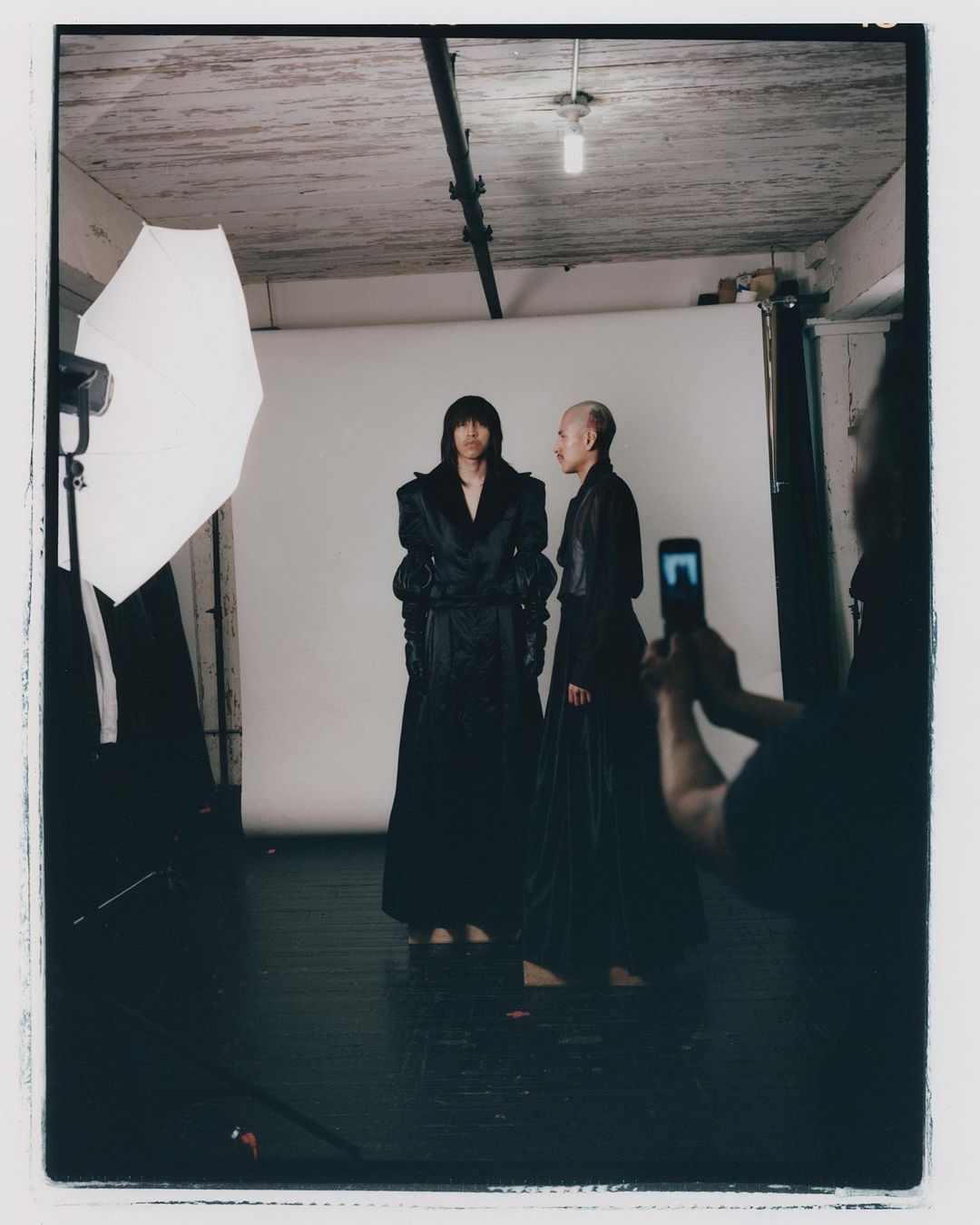 Two adult models pose in long black dresses in a studio setting. One person films the other with a smartphone.