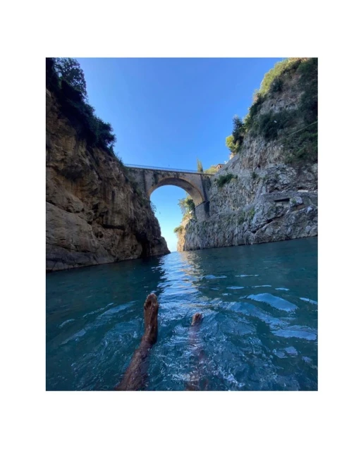 Person relaxing in turquoise water in a rocky gorge with a stone bridge overhead against a blue sky in Amalfi Coast, Italy.