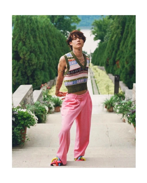 Stylish adult male poses on steps in a Ganni knit vest and pink trousers, with greenery and a building in the background on a sunny day.