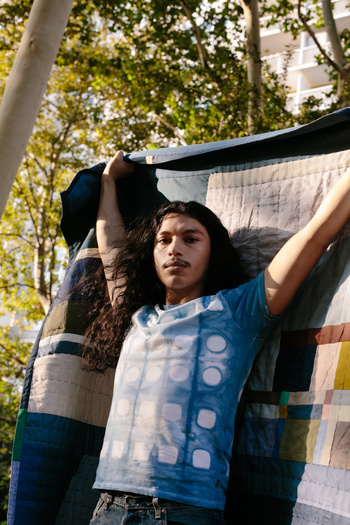 Young adult with long dark hair stands holding a colorful patchwork quilt outside on a sunny day in an urban park setting.