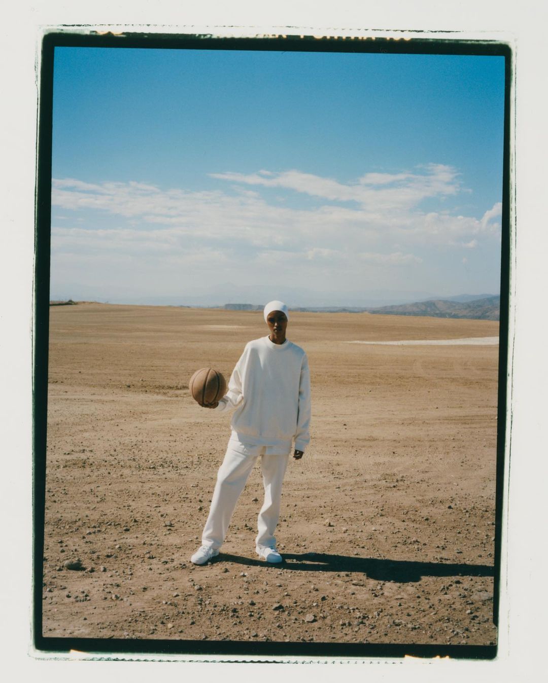 Young adult in white apparel stands in a desert landscape holding a basketball under a sunny blue sky with distant hills.