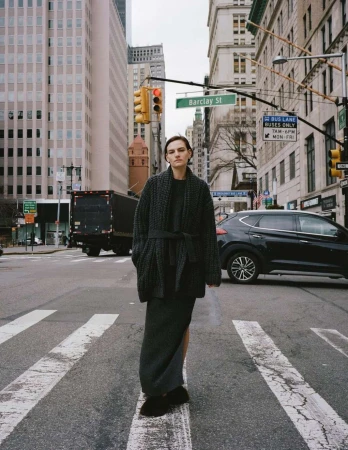 A woman stands on a city crosswalk in front of tall buildings, wearing a grey, belted cardigan and long skirt with city traffic behind.