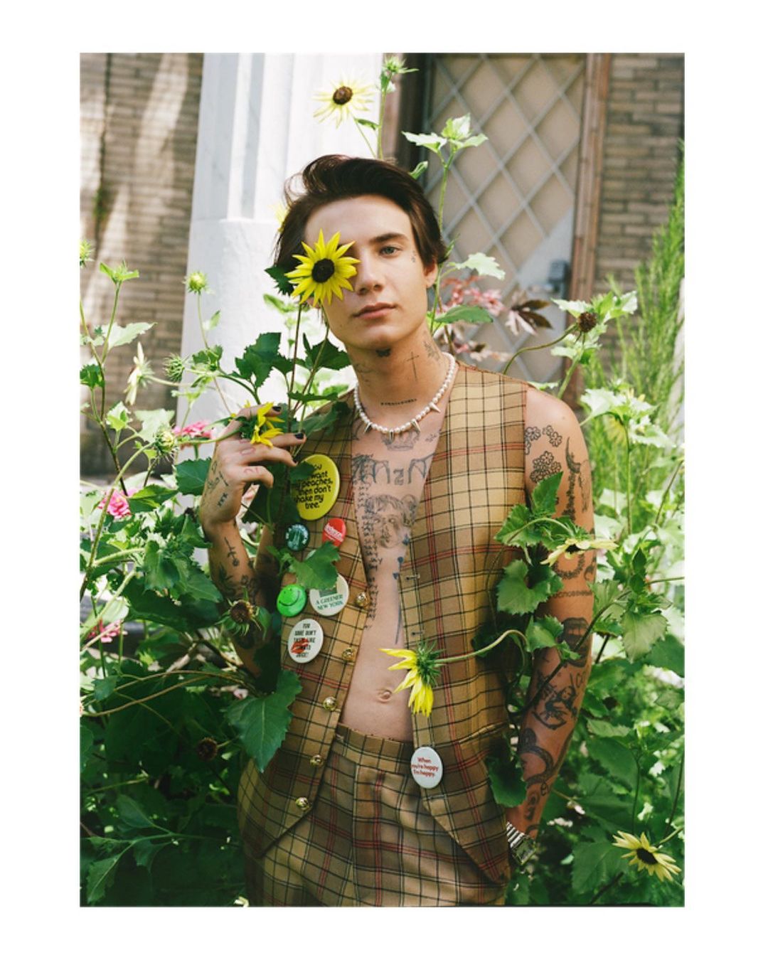 A young adult man with tattoos, wearing a vest and pearl necklace, posing with sunflowers in an outdoor garden setting with natural light.