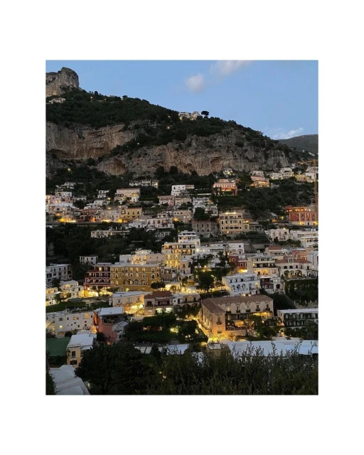 Picturesque Italian hillside town at dusk features tightly packed buildings on a slope under a mountain and a calming blue evening sky.
