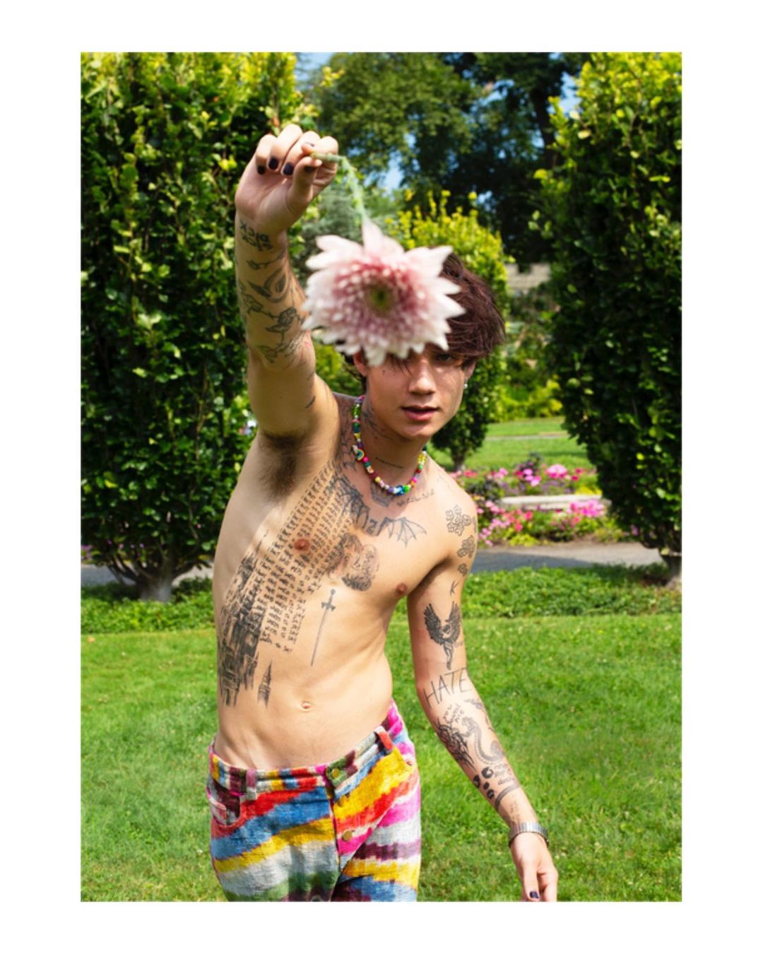 Young adult poses shirtless outdoors, holding pink dahlia flower. Wearing colorful pants, bead necklace, and arm tattoos in a garden setting.