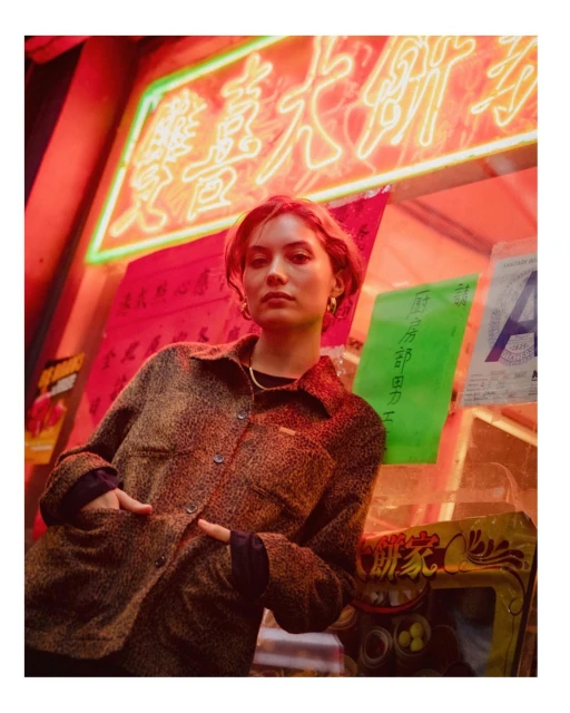 A young woman in a brown jacket poses against an urban backdrop with bright neon lights and a colorful store sign in a medium shot.