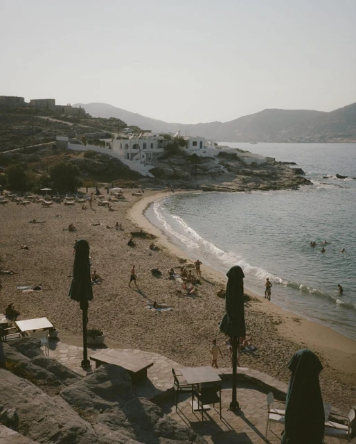 Scenic view of a sandy beach with people relaxing and swimming, featuring tables and umbrellas in the foreground and coastal buildings in the background.