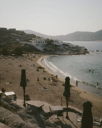 Scenic view of a sandy beach with people relaxing and swimming, featuring tables and umbrellas in the foreground and coastal buildings in the background.