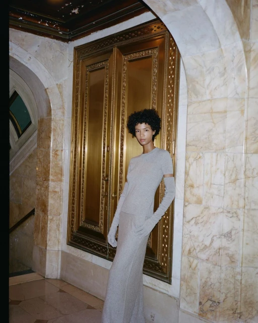A woman with afro hairstyle poses in a grey knit top and long skirt with gloves in a marble interior with ornate gold door frames.