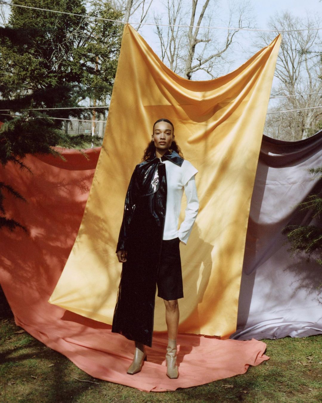 Elegant woman in avant-garde fashion posing outdoors against draped fabrics, showcasing a unique blend of textures and neutral tones.