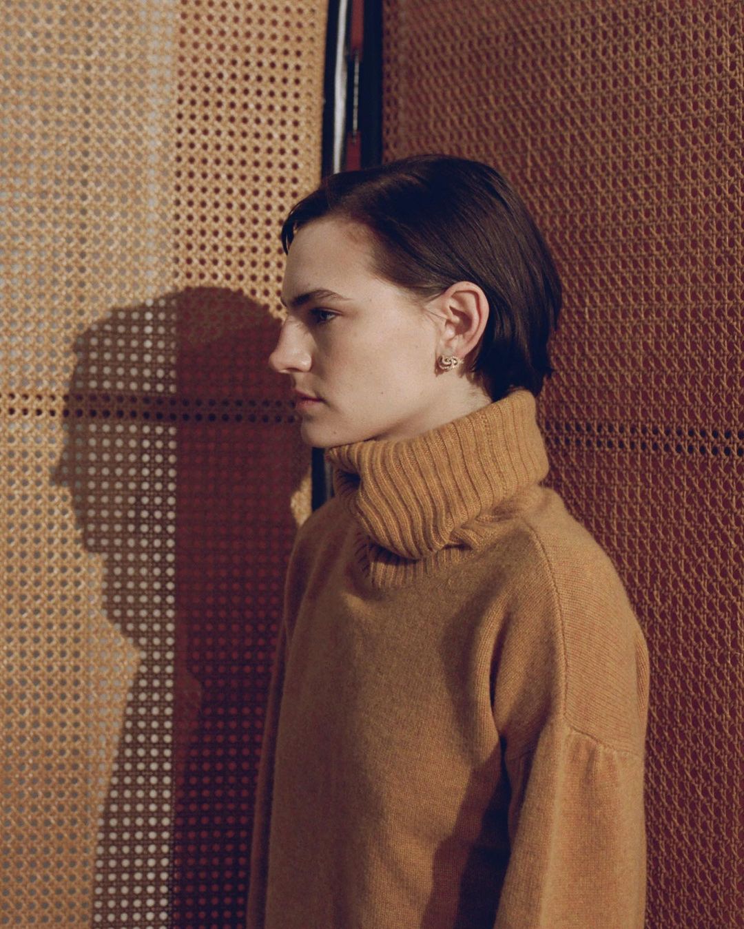 Profile portrait of a young adult woman with short brown hair wearing a brown turtleneck sweater, standing in front of a textured wall.