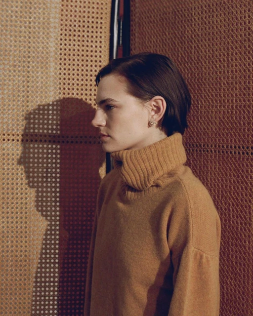 Profile portrait of a young adult woman with short brown hair wearing a brown turtleneck sweater, standing in front of a textured wall.