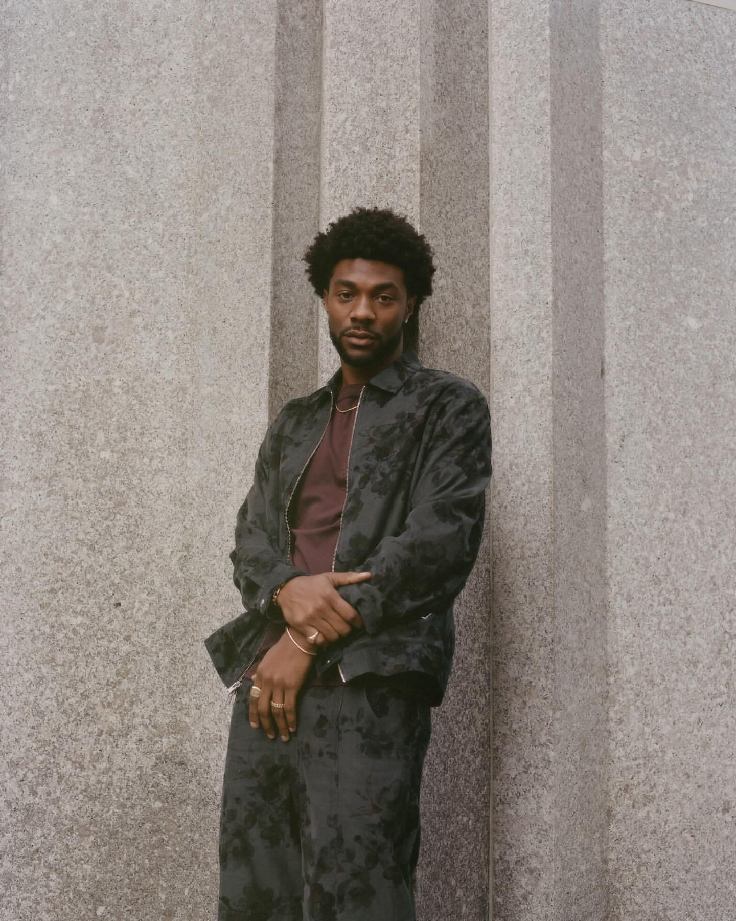 Black man with short dark hair wearing a patterned dark jacket and matching trousers posing against a grey stone column.