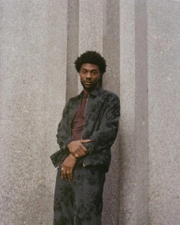 Black man with short dark hair wearing a patterned dark jacket and matching trousers posing against a grey stone column.