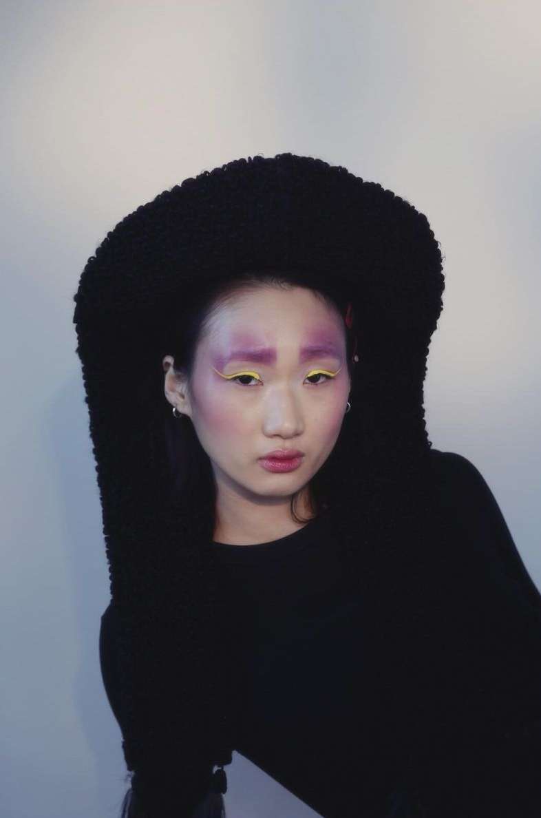 Asian woman with creative makeup poses in a black hooded outfit against a white background in a medium shot for a beauty concept.