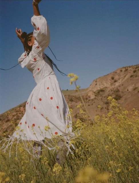 A woman in a white dress with red polka dots jumps in a field of yellow wildflowers against a mountain and blue sky.