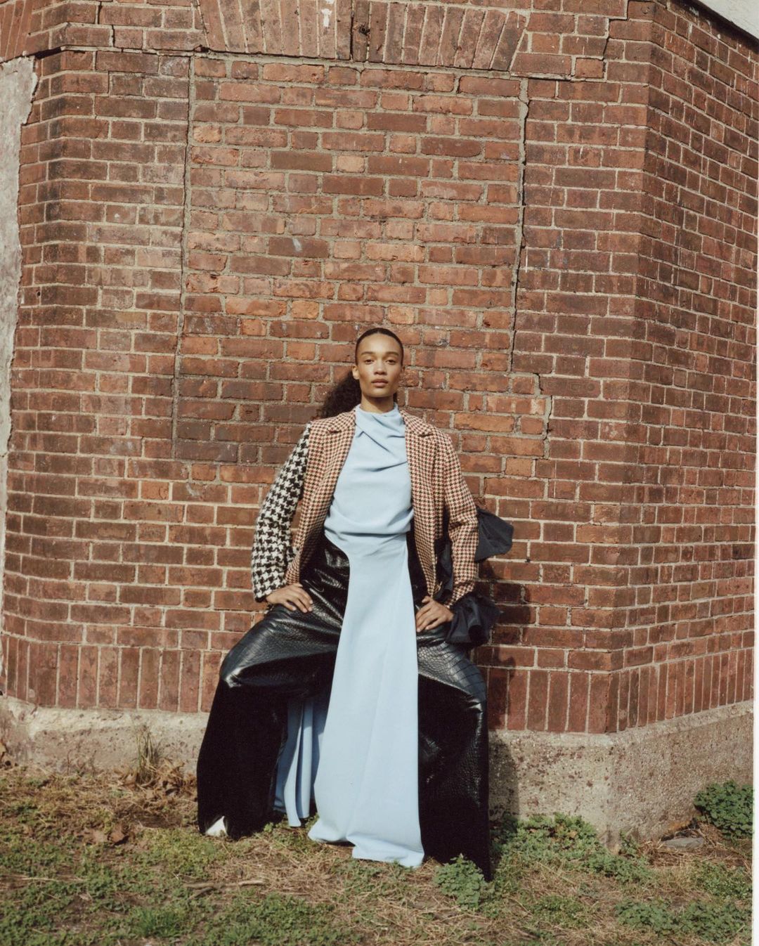 A woman in a blazer, light blue top, and wide, glossy pants poses against a brick wall, set in a grassy outdoor environment.