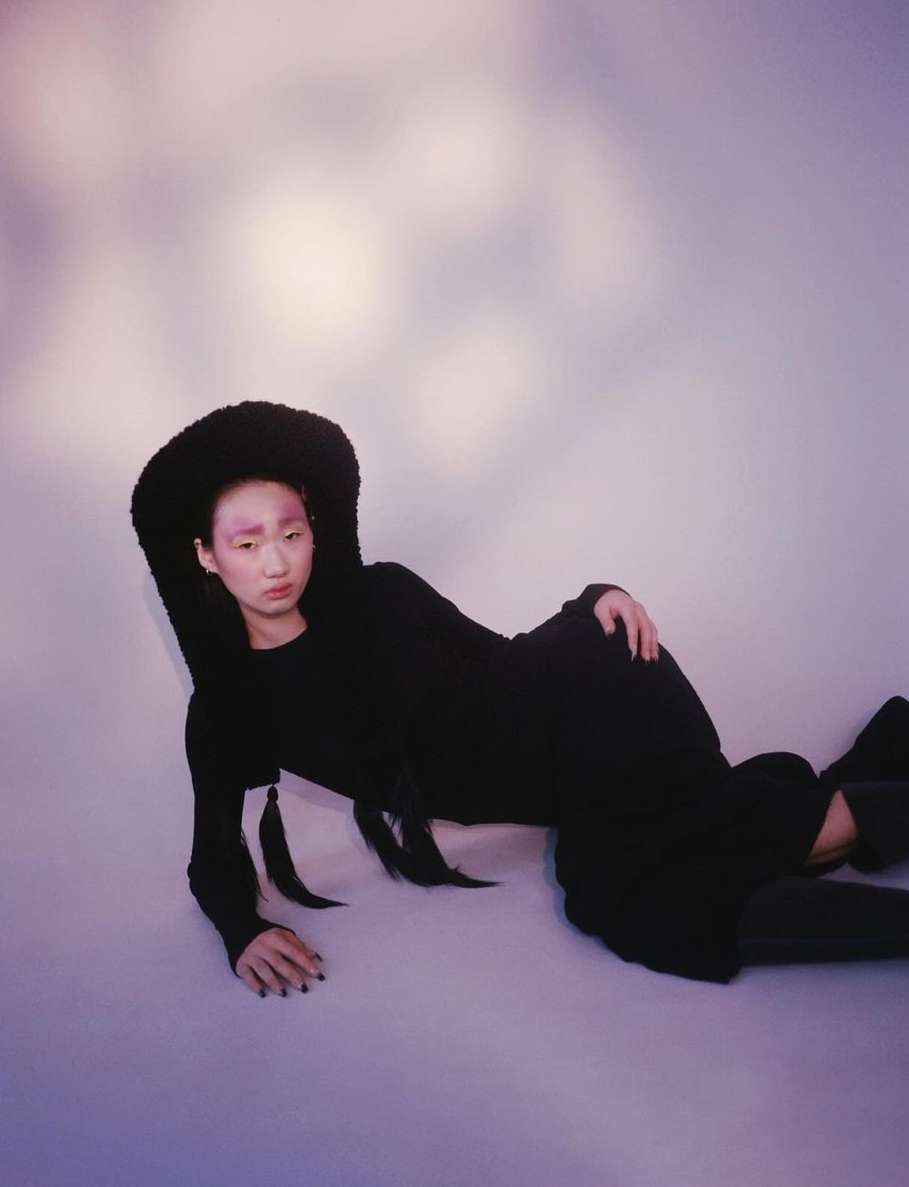 Asian woman in black dress and fluffy hat lies reclining against a cloud-like background, gazing at the camera with a neutral expression.