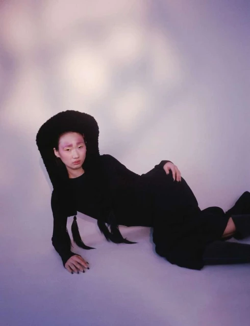 Asian woman in black dress and fluffy hat lies reclining against a cloud-like background, gazing at the camera with a neutral expression.