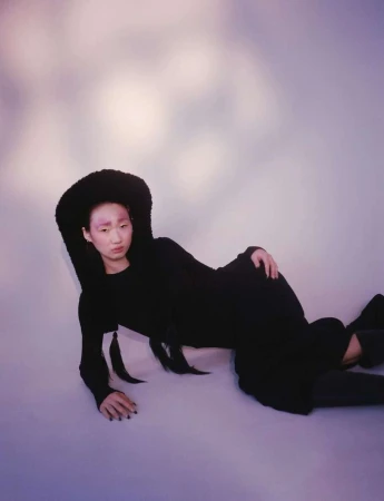 Asian woman in black dress and fluffy hat lies reclining against a cloud-like background, gazing at the camera with a neutral expression.