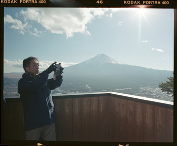 Adult male photographs Mount Fuji in natural light against a clear blue sky, standing on a wooden deck with visible film borders.