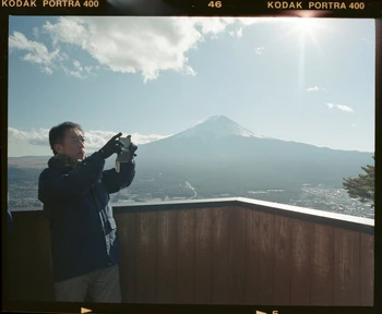 Adult male photographs Mount Fuji in natural light against a clear blue sky, standing on a wooden deck with visible film borders.