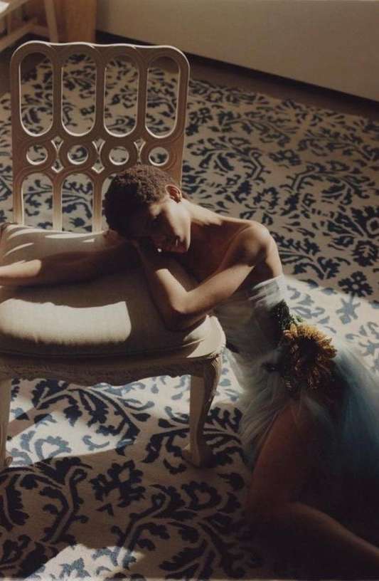 Woman in a light blue dress and floral accent, resting on a white decorative chair, on a blue and white patterned rug in sunlight.