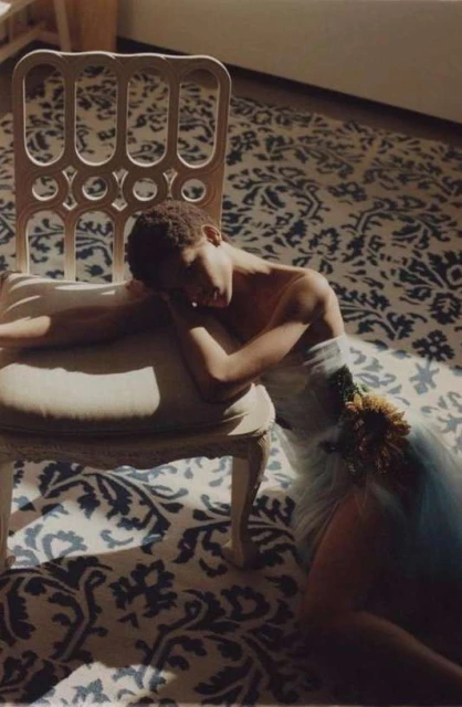 Woman in a light blue dress and floral accent, resting on a white decorative chair, on a blue and white patterned rug in sunlight.